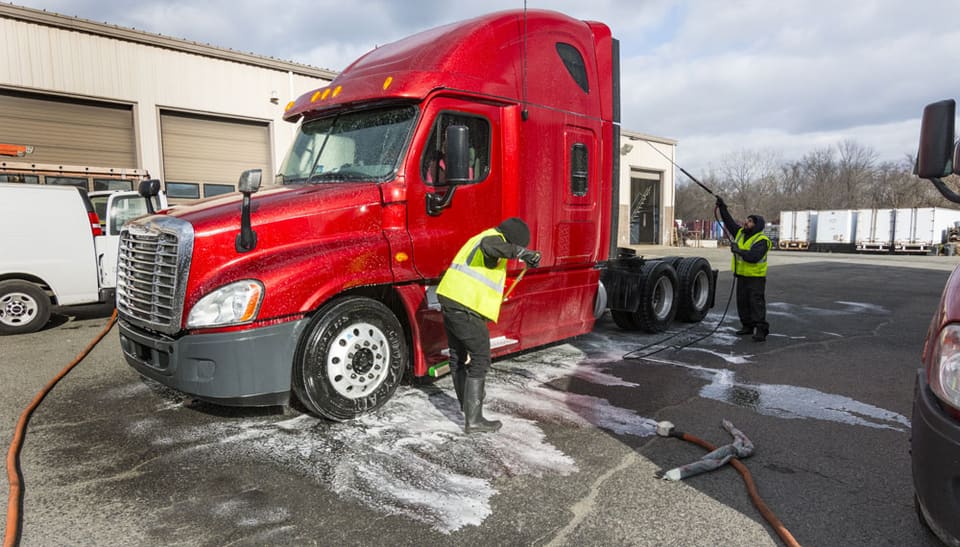 A red Freightliner semi truck covered in foam being washed by two technicians